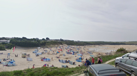 The image shows a wide sandy beach filled with groups of people sitting close to colourful windbreaks spread across the sand. Some people are walking along the shoreline while others are sitting or lying down on towels and beach chairs. There are a few small tents and beach shelters scattered around.