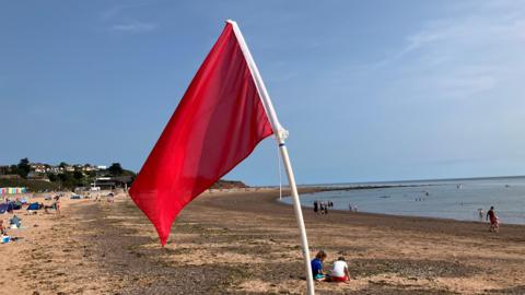 A red flag is blowing in the wind on a beach. Beachgoers are in the distance. Swimmers are in the water. The sky is blue.