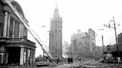 A black and white photograph showing the aftermath of the bombing on 7 May with the tower of the Prudential Assurance building tower surrounding by bombed out buildings, smoke and fire crews.