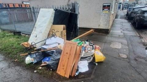 The picture is taken on the corner between two roads, with a pile of rubbished dumped. There is a white mattress, and some wooden draws in the pile. In the background there is a road leading up to a row of houses on the left. 