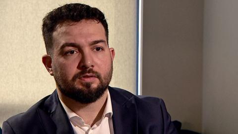 A man in a white shirt and black suit jacket sitting in a darkened room with the blind drawn. He has dark short hair and a dark beard.