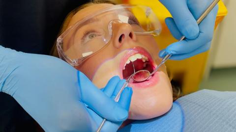 A woman lies back in a dentist chair with her mouth open. She is wearing protective glasses with the dentist examining her teeth with a mirror and dental probe 