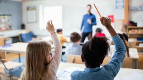 Two girls in classroom with raised hands, other pupils and a male teacher are visible in the background