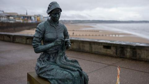The bronze sculpture is of a woman sitting on the plinth of the sculpture knitting. The bronze has weathered to a bluey black colour. The beach at Bridlington can be seen in the background.