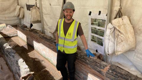 A person in safety gear stands inside an excavation area next to exposed brickwork and a narrow trench.The site is covered by a protective tent, with tools, bags, and a reference poster displaying images pinned to the wall. Bright sunlight filters in, highlighting the detailed layers of historic-looking masonry and the working environment.