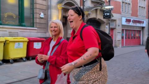 Two women walking and laughing in the street. One has short, straight, blonde hair and is wearing blue jeans and a red jacket. The other has black hair tied back and is wearing a red t-shirt and leopard print bottoms. 