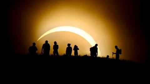A silhouetted image of the solar eclipse shows eight people standing on a hill watching the eclipse. Only a sliver of sun is visible and it appears to be very large against the silhouettes of the people.