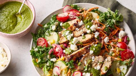 A white plate of kale leaves topped with sliced chicken and vegetables with a small bowl of green sauce on a stone surface