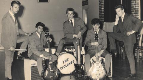 Five young men photograhped in the 1950s wearing suits typical of that period. Two are holding guitars, one is standing over a drumkit with "The Cyclones" written on the front.
