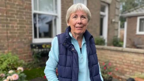 A woman wearing a blue gilet is looking into the camera and standing in front of the window of a house. She has short grey hair and is wearing a turquoise quarter-zip long-sleeve top.