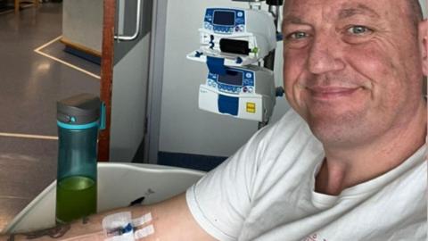 A selfie taken by a man with very short hair wearing a white t-shirt. He is smiling at the camera. Behind him is a small machine and he has an IV line taped onto his arm. Plain walls and floors show he is in a hospital.