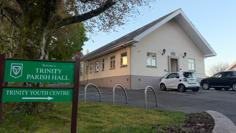 Exterior view of Trinity Parish Hall, a single‑storey cream building with parked cars, beside a green sign reading “Welcome to Trinity Parish Hall” and “Trinity Youth Centre,” under a large tree.