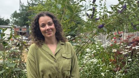 A woman with long brown hair and a green top stood in a garden