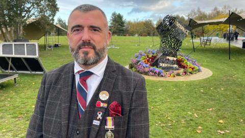 A man with short hair and a beard, wearing a grey checked suit with a red, white and blue tie, pride and remembrance badges and a military medal, stands in front of the LGBT+ armed forces memorial, which resembles an unravelling piece of paper and is surrounded by brightly coloured flowers.