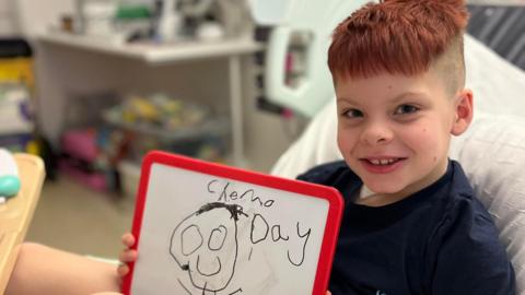 Jasper Hodgson smiles at the camera, he holds a whiteboard drawing reading "chemo day". 