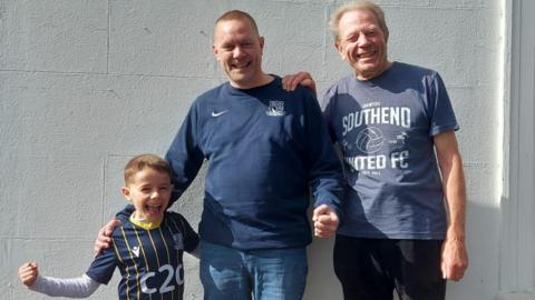 A boy with a Southend United shirt, a man with a Southend United jumper, and an older man with a Southend United T-shirt. All of them have their arms round each other.