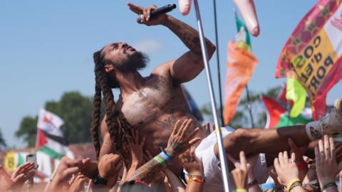 A performer with long dreadlocks sings into a microphone while crowd surfing.