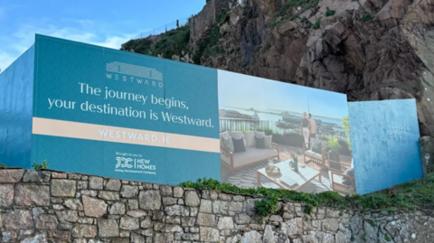The image shows large blue advertising hoarding for the Westward residential development mounted against a rocky cliff above a stone wall. The sign includes promotional text, the website Westward.je, and an image of an outdoor terrace overlooking the water.