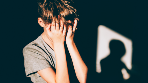 A boy with brown hair and a grey tshirt with his face in his hands in front of a black background