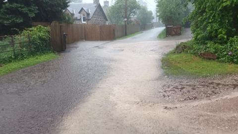 Muddy water flows down a rural road. The road is flanked by green hedges and one side of the road is a traditional, stone-built house with a peaked slate roof.