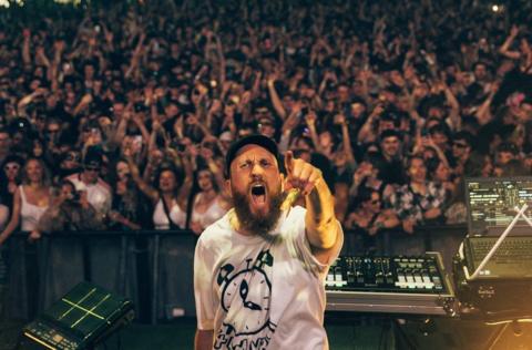 A man with a beard and black cap points at the camera as he DJS in front of a large crowd at Love Saves The Day in Bristol
