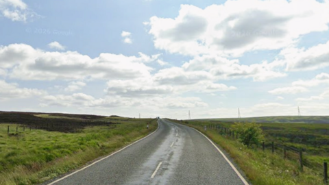 A road running up to the horizon with moorland on either side. Above is a blue sky with some clouds. Power lines can be seen on the horizon in the distance.