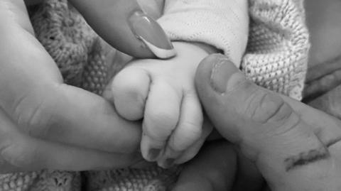A baby's hand, held by a woman's hand and a man's hand. It is a greyscale image.