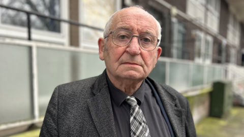 Lord John Bird wearing glasses and a grey jacket over a black shirt and black and white tie. He is standing in front of a black of flats