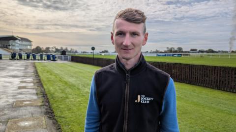 Tom Ryall is standing in the middle of the shot, wearing a blue jumper and black fleece vest on top. He is looking at the camera with a neutral expression. He is standing in front of the racetrack and the stands.