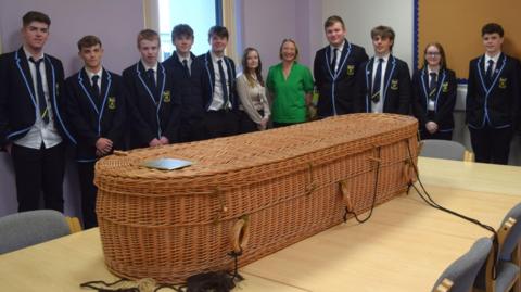Moffat Academy sixth year pupils along with teachers and people involved in the course stand in a classroom. The pupils are wearing school blazers. On a table in front of the group is an empty wicker coffin.