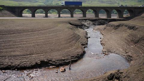 Dried up reservoir bed with only a narrow stream of water flowing through. There is a bridge in the background with a blue lorry on it. 