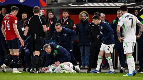 Sam Szmodics of Republic of Ireland is helped by Republic of Ireland head coach Heimir Hallgrimsson after a head injury. Szmodics is wearing an all white Republic kit and laying on the ground