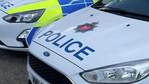 The bonnets of two Manx police cars. They are white and have the Isle of Man Constabulary crest above the word 'police' in blue lettering.  The side of the car furthest away is blue and yellow.