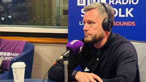 A man is sat in front of a purple microphone and wearing headphones inside a radio studio
