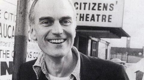 A black and white shot of a man outsizxe the Citizens' Theatre in Glasgow. He is clean shaven with short hair and has a wide smile. He wears a dark blazer, a shirt unbuttoned at the neck and a light t-shirt, with a Citizens' Theatre badge on his lapel.