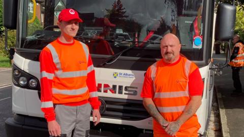 Two refuse collectors dressed in orange hi-vis tops lean on the front of a white bin lorry. Dan Gardiner is bald with a goatee. Matko Spondreht is taller and has a orange cap on.