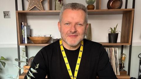 A man in a black long sleeve top wearing a yellow lanyard sits at a table looking at the camera.