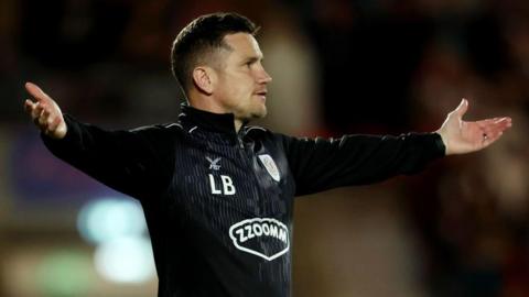 Crewe Alexandra manager Lee Bell is standing on the touchline with his arms outstretched