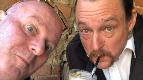 Selfie of Steve Lilley, who is on the left, who has a bald head with Steve Marriott on the right, who has dark hair, a moustache, am earring and is drinking from a straw