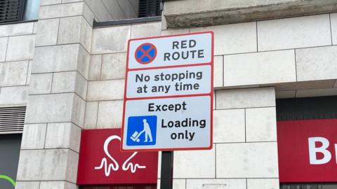 A white road sign with red borders on a street in Brighton, it reads "red route, no stopping at any time, except loading only"