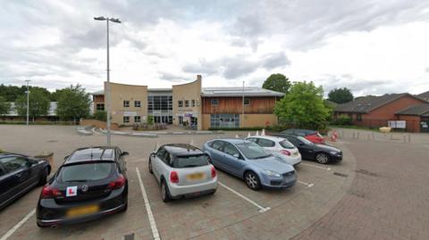 Line of cars parked in a car park with a community centre that is made from pale stone and wood behind them 