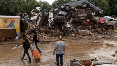 A man stands and stares at a pile of cars that have been damaged in floods. The cars have been moved from the road