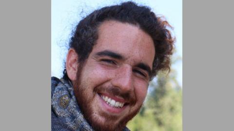 Adam Cornell smiling at the camera. He has curly reddish brown hair, freckles, a beard and a moustache. He is wearing a blue scarf. In the background there are trees and sky.