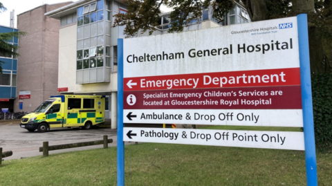 An exterior shot of a sign at Cheltenham General Hospital showing the name of the hospital and points towards the emergency department, an ambulance is parked by the entrance to the left