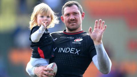 Alex Goode waves to fans while holding his daughter
