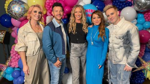 A group of five people smiling and standing next to each other in front of a blue, yellow, pink and silver balloon feature wall.