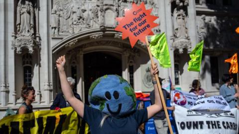 A woman with a large Planet Earth mask obscuring her face celebrates while holding a sign saying "The fight for our future" outside the High Court in London. Protesters in the background also hold signs and banners