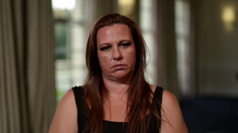 A woman wearing a black tank top and long reddish brown hair sits and stares at the camera.