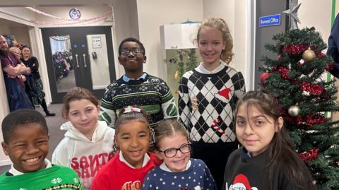 Seven children stand in front of a Christmas tree at their primary school. The five girls and two boys are all wearing festive jumpers which are traditional colours of red, green, white and blue.