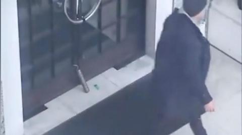 CCTV image showing a cylindrical object propped upright against the glass doors of a building entrance, with a small green lighter lying nearby, as a man in a dark coat walks away.